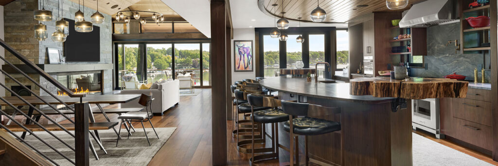 Midcentury-to-transitional lake home interior featuring an open great room and kitchen with wood ceilings, stone fireplace, modern lighting, and large windows overlooking the lake.